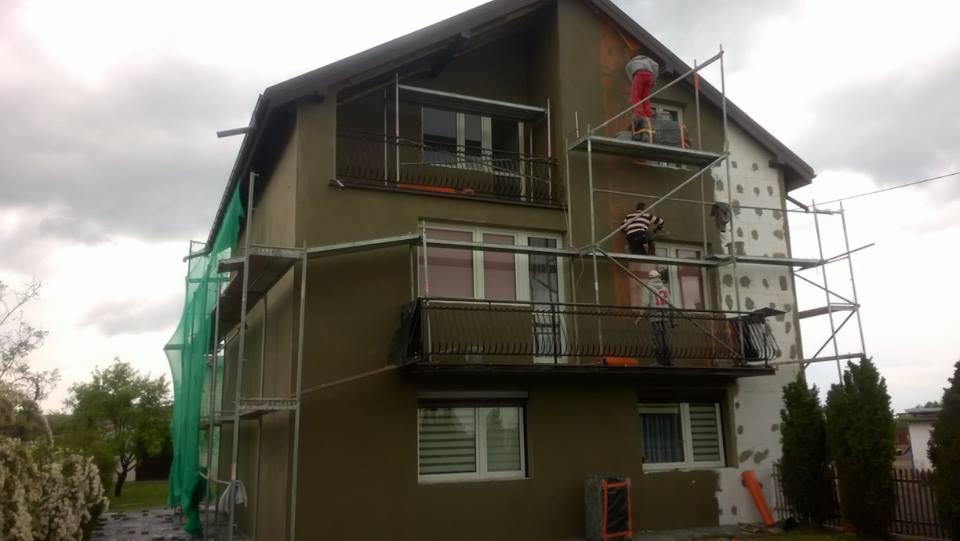 Dwupiętrowy dom w trakcie prac elewacyjnych: część ściany ocieplona styropianem, druga część z nałożonym tynkiem w kolorze oliwkowym; dwóch pracowników na rusztowaniu, jeden w czerwonych spodniach,...