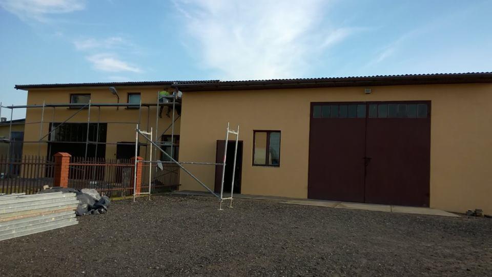 Ocieplony budynek gospodarczy z brązową bramą garażową i pracownikiem na rusztowaniu, widoczne materiały budowlane i ogrodzenie.