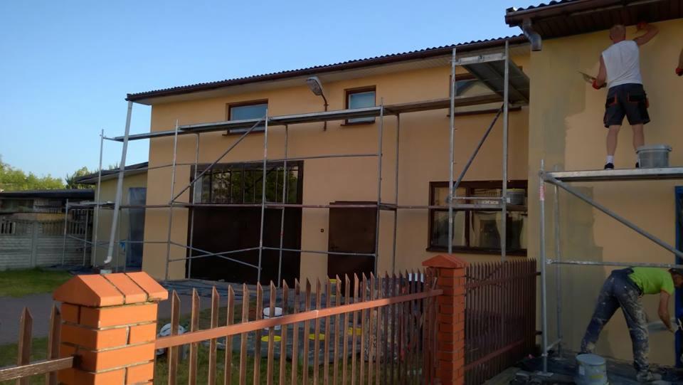 Dwóch pracowników na rusztowaniu podczas nakładania tynku na elewację domu jednorodzinnego w słoneczny dzień, widoczny fragment ogrodzenia z ceglanym słupkiem.