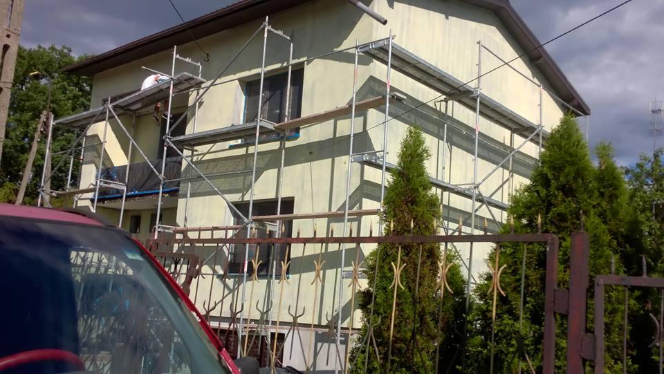Dom dwukondygnacyjny w trakcie prac elewacyjnych, widoczne rusztowania, fragment czerwonego samochodu i metalowe ogrodzenie na pierwszym planie.