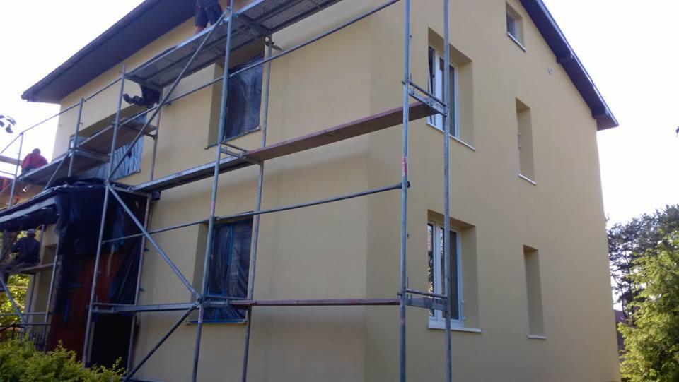Dwupiętrowy dom z jasną elewacją w trakcie prac wykończeniowych, widoczne rusztowania i pracownicy.