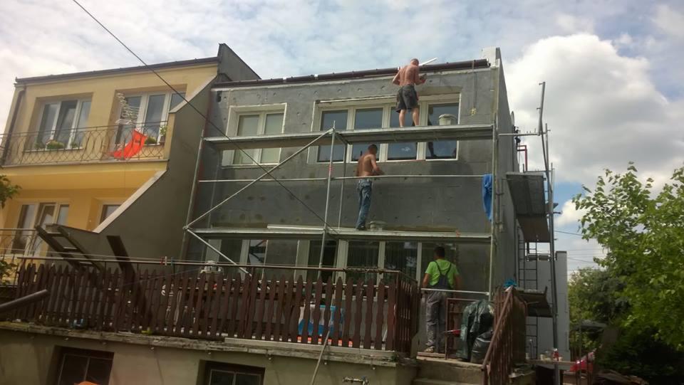 Trzech mężczyzn pracuje na rusztowaniu przy ocieplaniu ściany budynku, widoczne płyty izolacyjne i narzędzia, sąsiedni budynek z balkonem i suszącą się czerwoną koszulką.