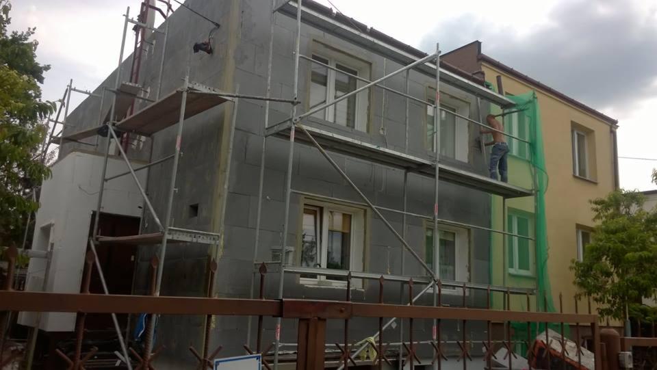 Dwukondygnacyjny dom w trakcie prac ociepleniowych, widoczne rusztowania, częściowo ocieplona ściana szarymi płytami styropianowymi, pracownik na rusztowaniu w jeansach i bez koszulki, sąsiedni...