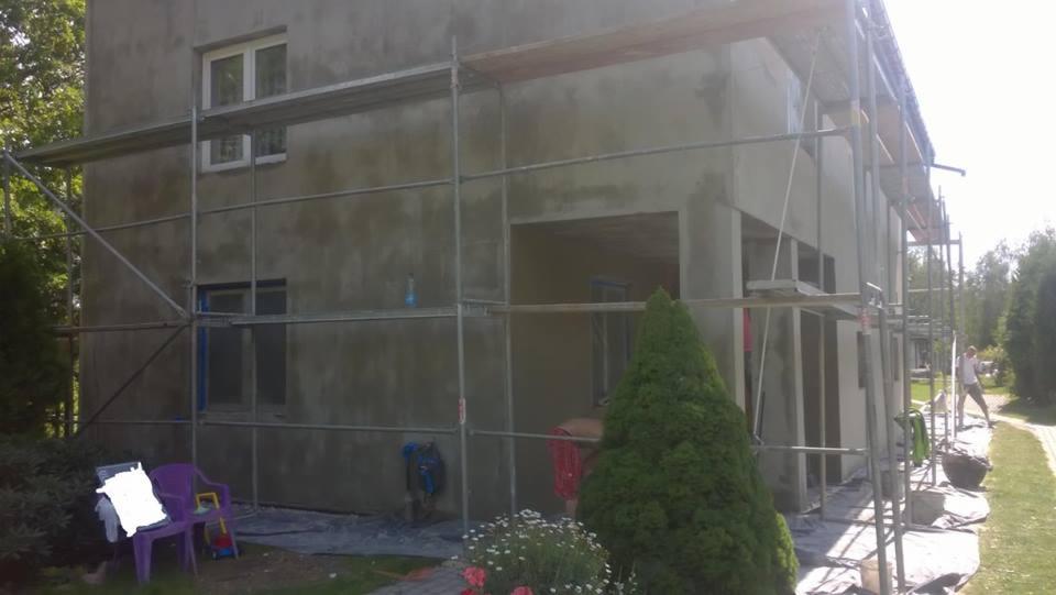 Dom w trakcie tynkowania elewacji, widoczne rusztowania, surowy tynk, zabezpieczone okna folią, w tle zieleń ogrodu, na pierwszym planie krzesła ogrodowe.