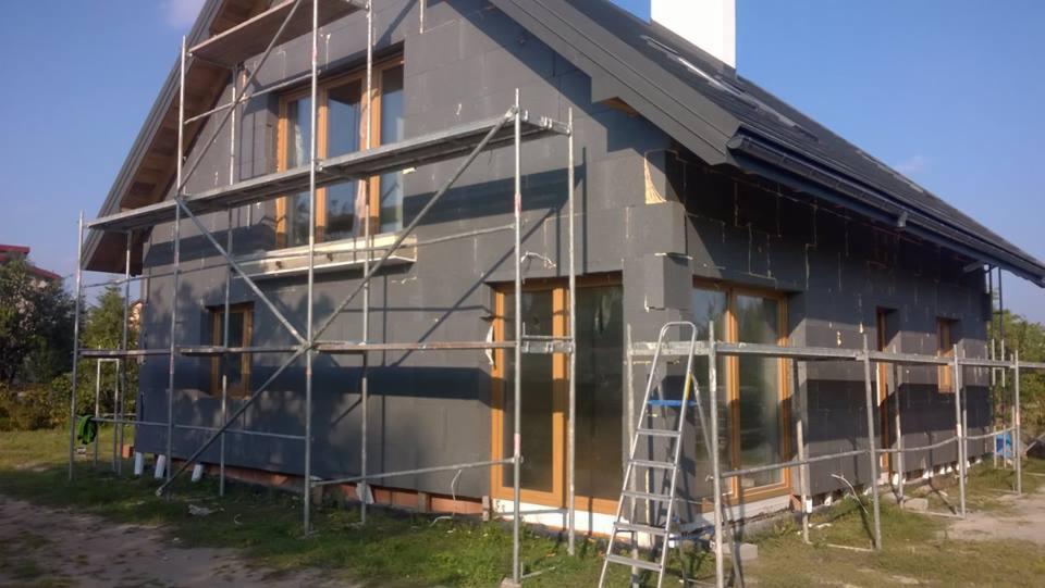 Dom jednorodzinny w trakcie ocieplania szarą wełną mineralną, widoczne rusztowanie i drabina oparta o ścianę, okna z drewnianą stolarką.
