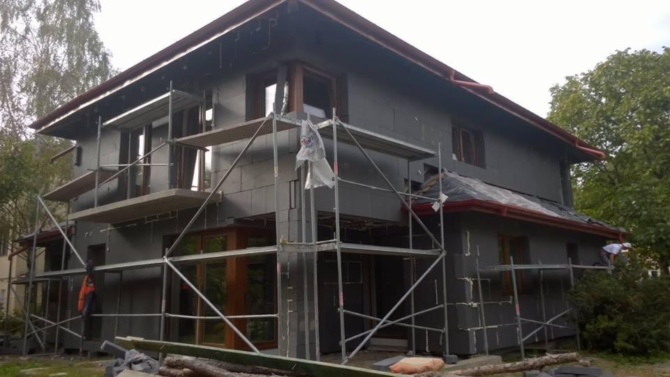 Dwukondygnacyjny dom w trakcie ocieplania elewacji, widoczne rusztowania, szare płyty izolacyjne na ścianach, pracownik w białej koszulce na rusztowaniu przy dachu.