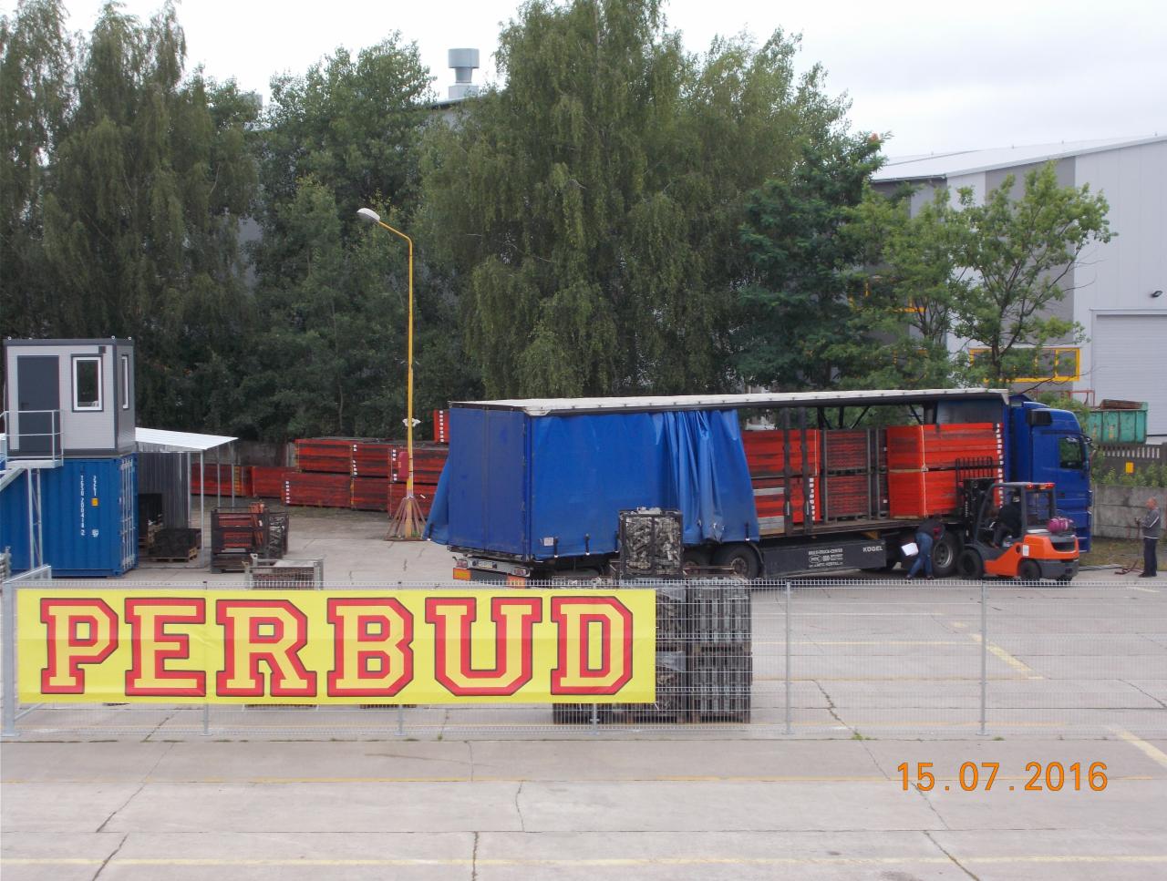 Ciężarówka z niebieską plandeką załadowana czerwonymi elementami szalunkowymi, obok wózek widłowy, w tle składowisko materiałów budowlanych i budynek przemysłowy, na pierwszym planie baner z nazwą...