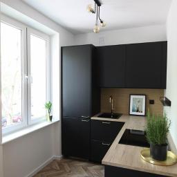 Korda Sp z o.o. - Nowoczesna, mała kuchnia z czarnymi szafkami, drewnianym blatem i podłogą w jodełkę, oświetlona lampą z trzema żarówkami Edison, z rośliną w doniczce na parapecie i ziołami w doniczce na blacie.