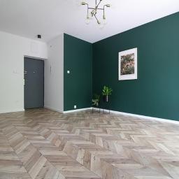 Korda Sp z o.o. - Wnętrze mieszkania po remoncie: parkiet w jodełkę, ściana pomalowana na ciemnozielono, nowoczesna lampa sufitowa, drzwi wejściowe w kolorze grafitowym.
