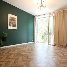 Korda Sp z o.o. - Pokój z parkietem ułożonym we wzór jodełki, ściana pomalowana na ciemnozielono, obraz w ramie, rośliny w doniczkach, balkon z otwartymi drzwiami balkonowymi i beżowa zasłona.