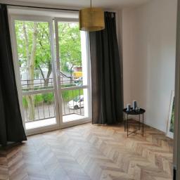 Korda Sp z o.o. - Salon w Warszawie po remoncie z parkietem ułożonym w jodełkę, widok na balkon i złota lampa wisząca.