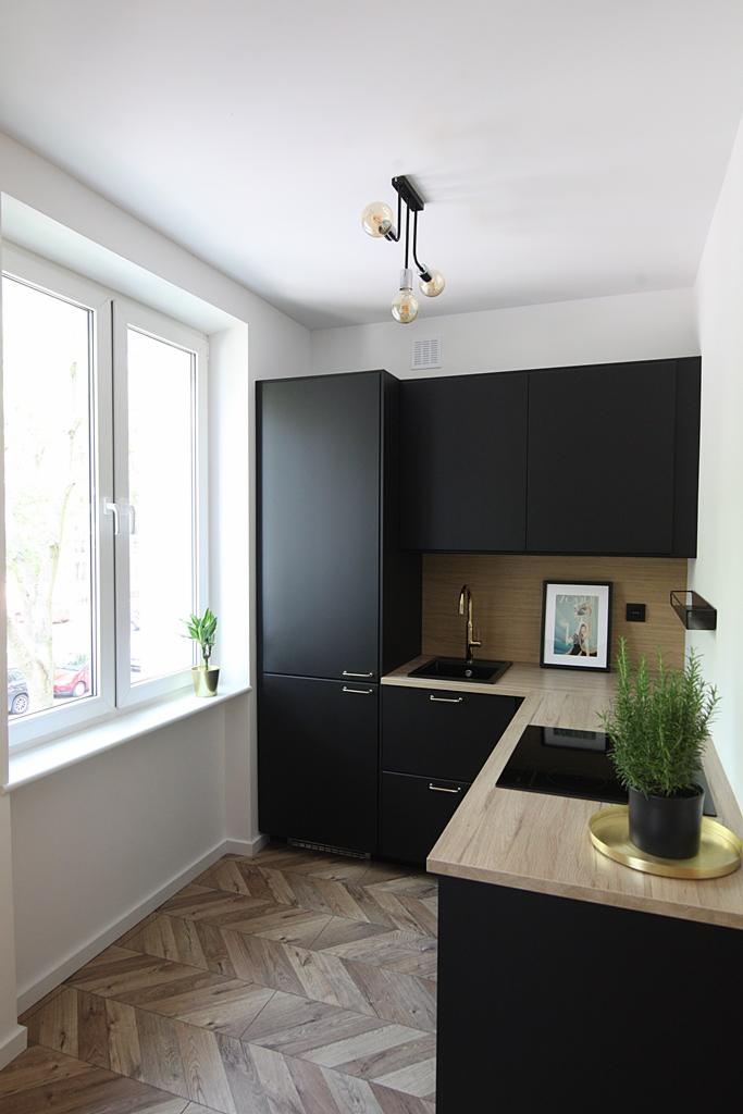 Nowoczesna, mała kuchnia z czarnymi szafkami, drewnianym blatem i podłogą w jodełkę, oświetlona lampą z trzema żarówkami Edison, z rośliną w doniczce na parapecie i ziołami w doniczce na blacie.