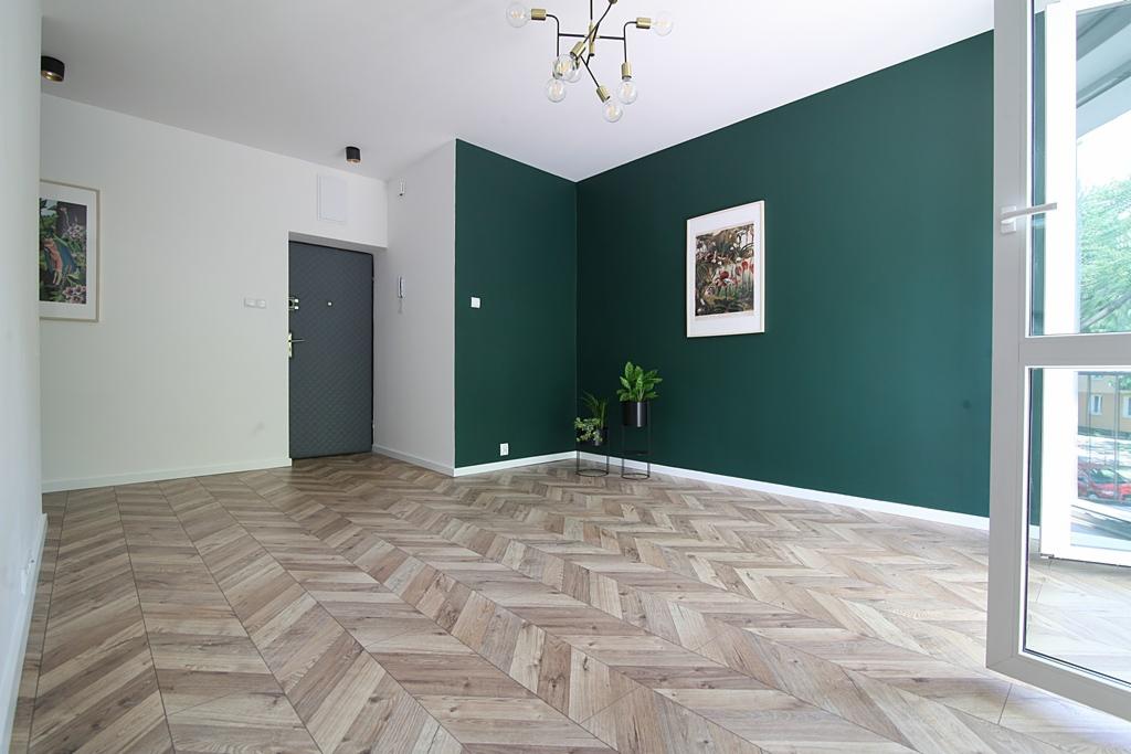 Wnętrze mieszkania po remoncie: parkiet w jodełkę, ściana pomalowana na ciemnozielono, nowoczesna lampa sufitowa, drzwi wejściowe w kolorze grafitowym.