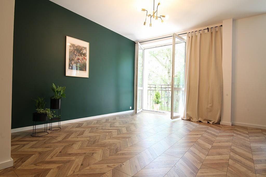 Pokój z parkietem ułożonym we wzór jodełki, ściana pomalowana na ciemnozielono, obraz w ramie, rośliny w doniczkach, balkon z otwartymi drzwiami balkonowymi i beżowa zasłona.