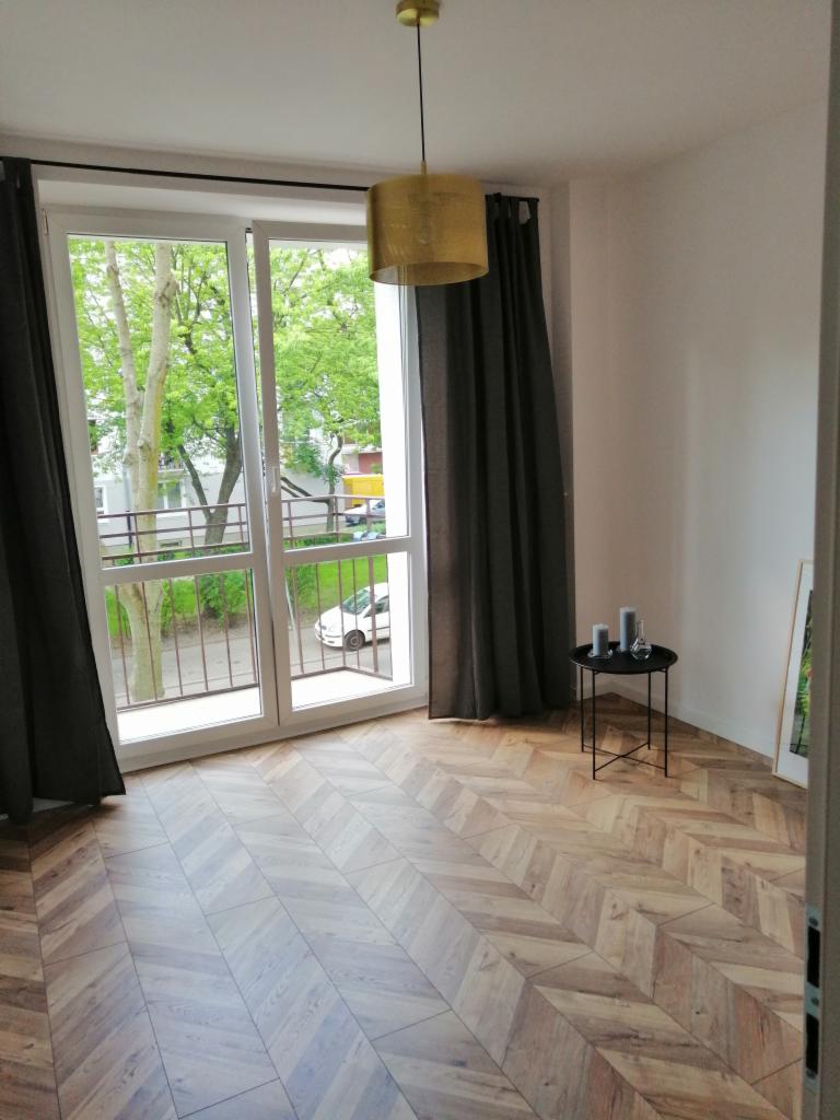 Salon w Warszawie po remoncie z parkietem ułożonym w jodełkę, widok na balkon i złota lampa wisząca.