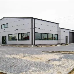 Zewnętrzny widok ukończonej hali magazynowej z szarej blachy trapezowej, z widocznymi oknami, drzwiami i utwardzonym placem manewrowym przed budynkiem.