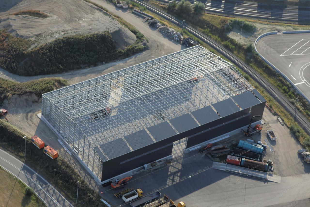 Widok z góry na budowaną halę z metalową konstrukcją szkieletową, częściowo obłożoną szarymi panelami, z widocznymi maszynami budowlanymi i elementami konstrukcyjnymi na placu budowy.
