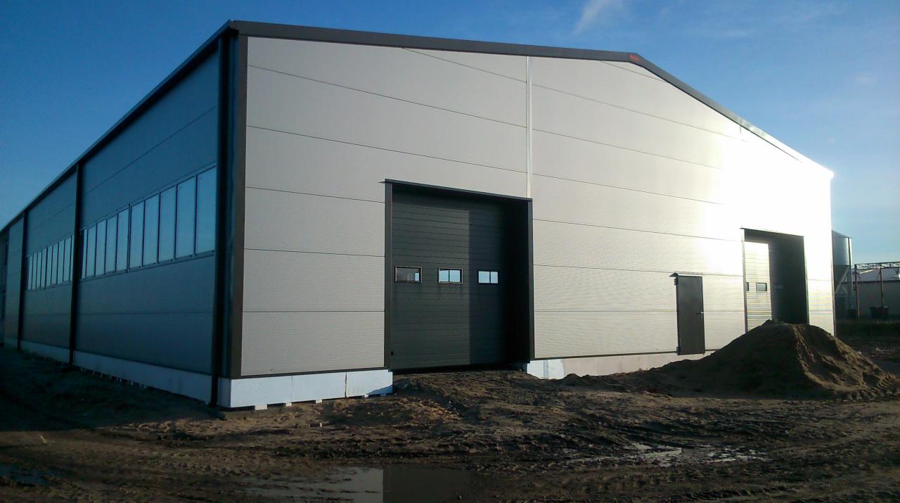 Nowoczesna hala magazynowa z szarej blachy trapezowej, widoczne okna wzdłuż boku i duże, ciemne wrota wjazdowe, teren wokół hali częściowo utwardzony, z widoczną pryzmą ziemi.