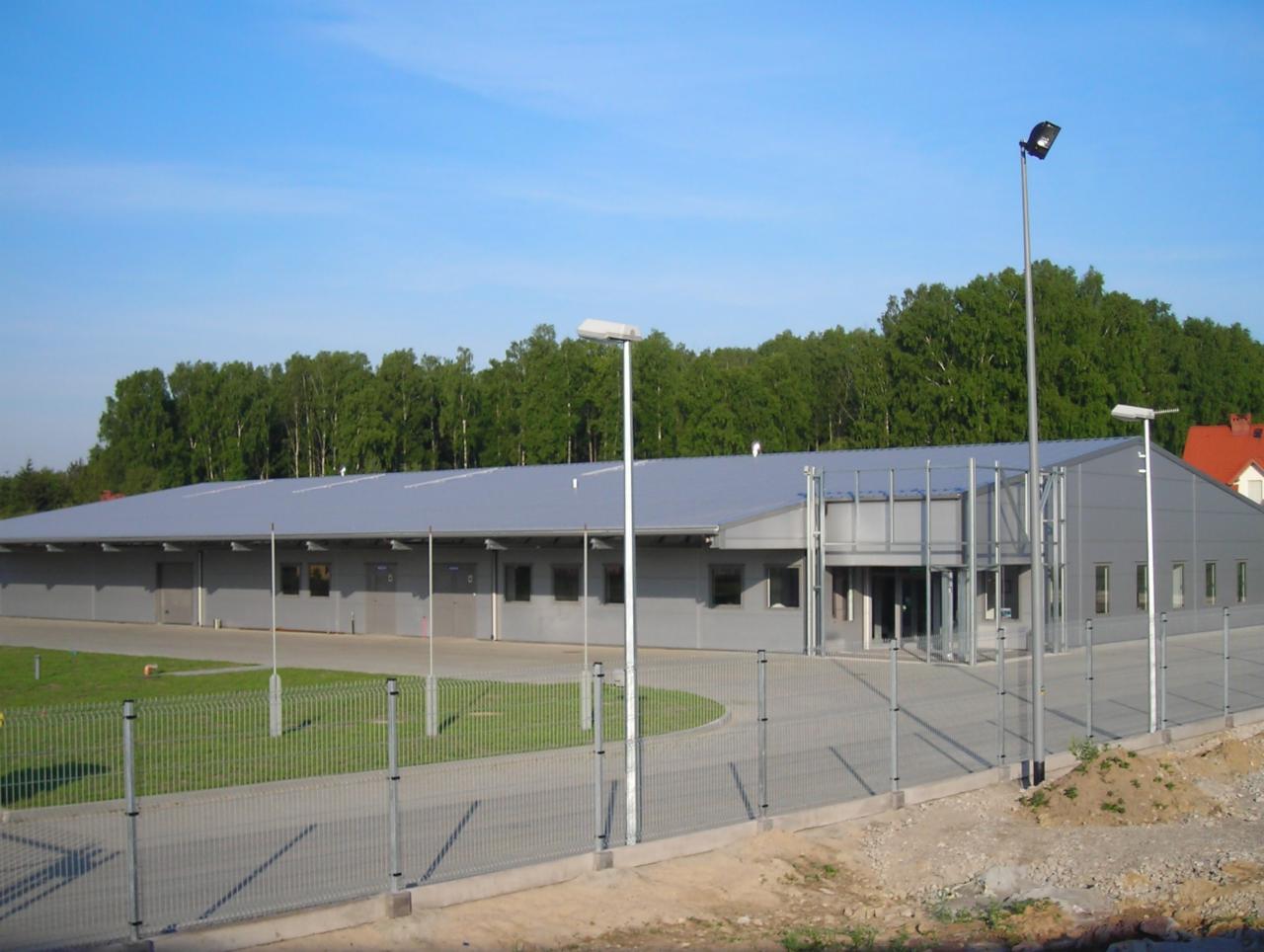 Gotowa hala przemysłowa z szarej blachy trapezowej, widoczna z zewnątrz, ogrodzona metalową siatką, z zielonym trawnikiem i drzewami w tle.