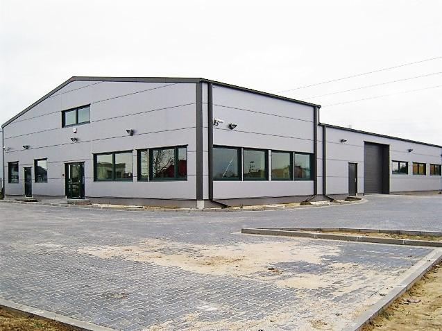 Zewnętrzny widok ukończonej hali magazynowej z szarej blachy trapezowej, z widocznymi oknami, drzwiami i utwardzonym placem manewrowym przed budynkiem.