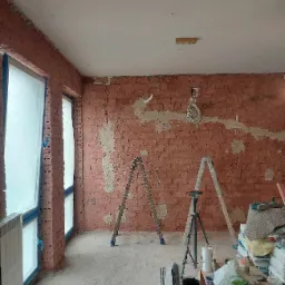 Ceglana ściana w trakcie przygotowania do gładzi, z widocznymi śladami po instalacjach. Okna zabezpieczone folią, drabiny i narzędzia w pomieszczeniu.