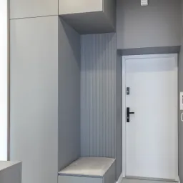 Szara zabudowa w przedpokoju z siedziskiem i szafkami, białe drzwi wejściowe, szare ściany i podłoga z płytek. Minimalistyczny design.