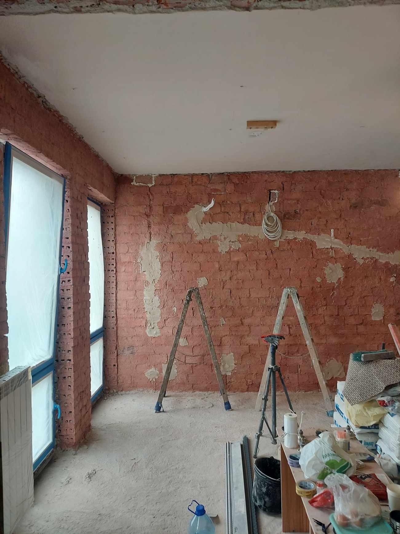 Ceglana ściana w trakcie przygotowania do gładzi, z widocznymi śladami po instalacjach. Okna zabezpieczone folią, drabiny i narzędzia w pomieszczeniu.