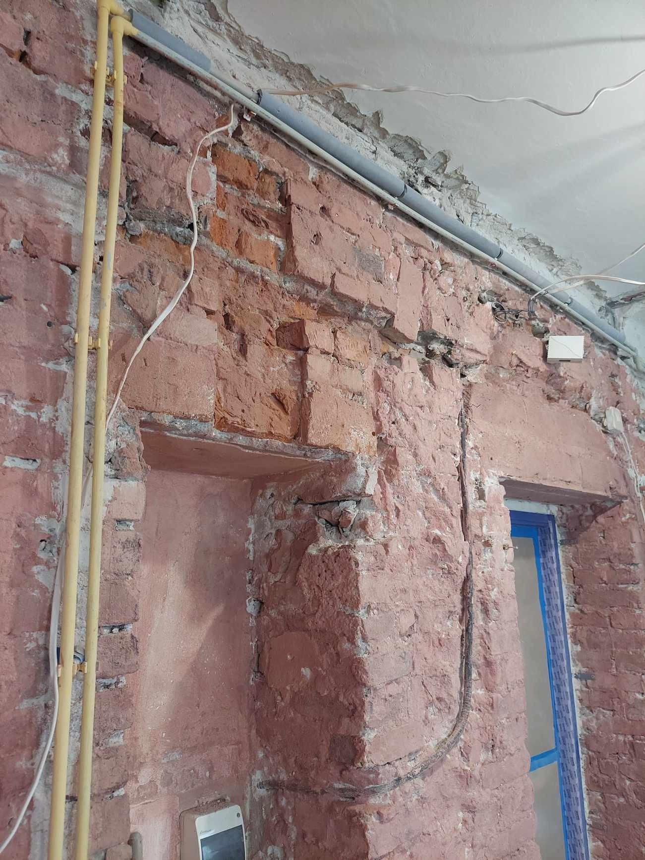 Odsłonięta ceglana ściana z elementami instalacji elektrycznej i rur. Widoczne ślady po kuciu i naprawach. Fragment okna z niebieską ramą.