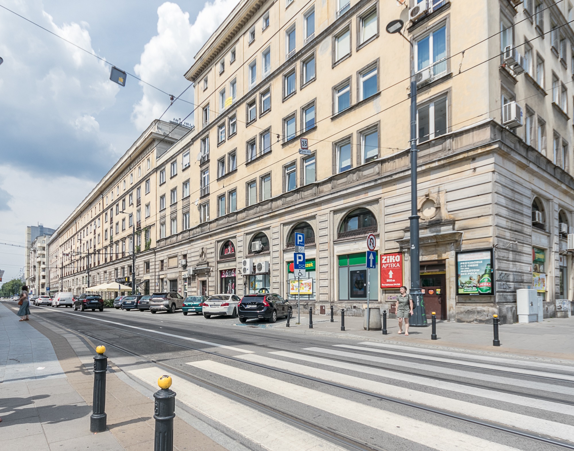 Elewacja kamienicy z oknami i klimatyzacją, widok z ulicy z przejściem dla pieszych i torami tramwajowymi. Na elewacji szyldy reklamowe i apteki.