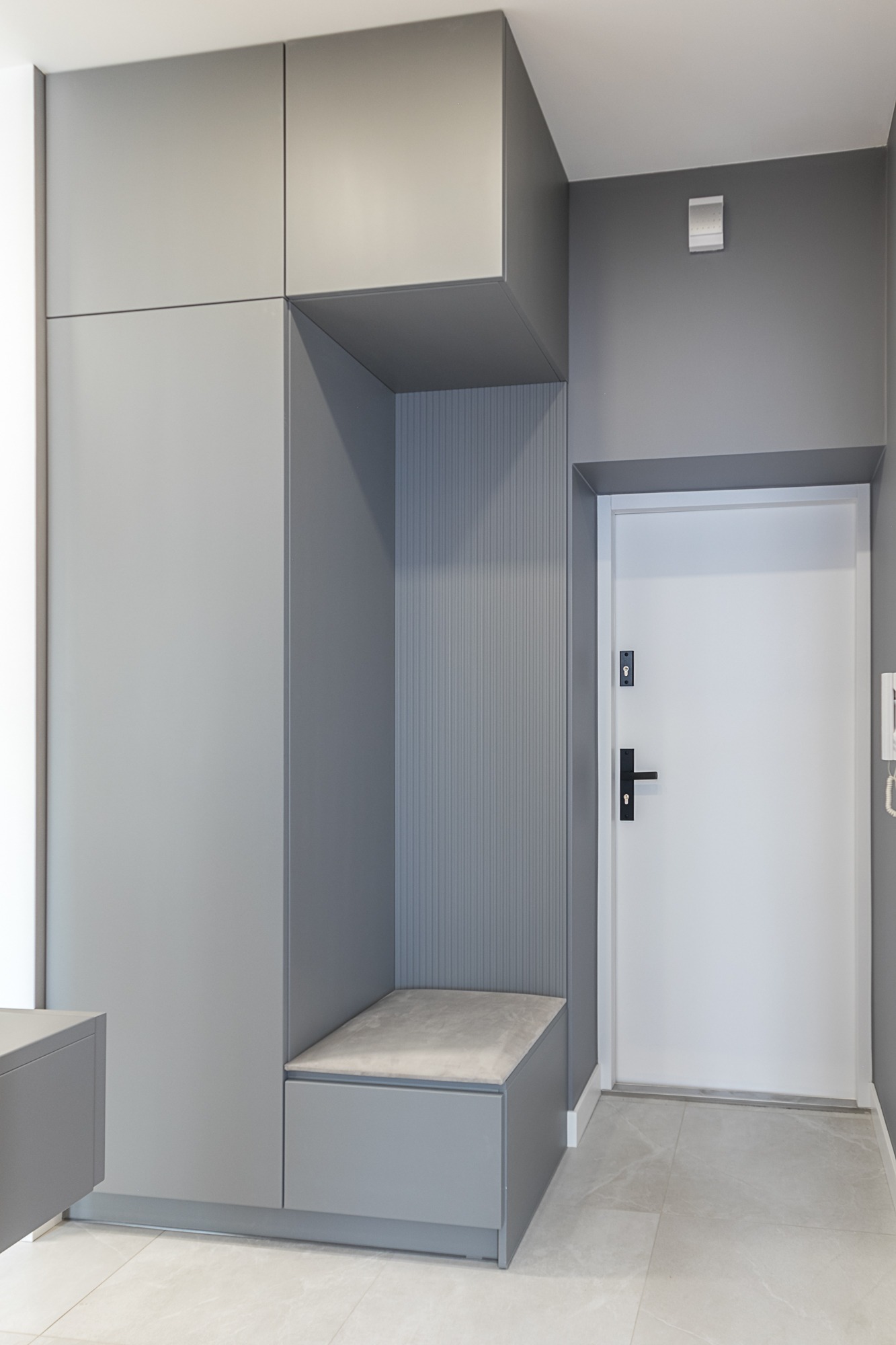 Szara zabudowa w przedpokoju z siedziskiem i szafkami, białe drzwi wejściowe, szare ściany i podłoga z płytek. Minimalistyczny design.