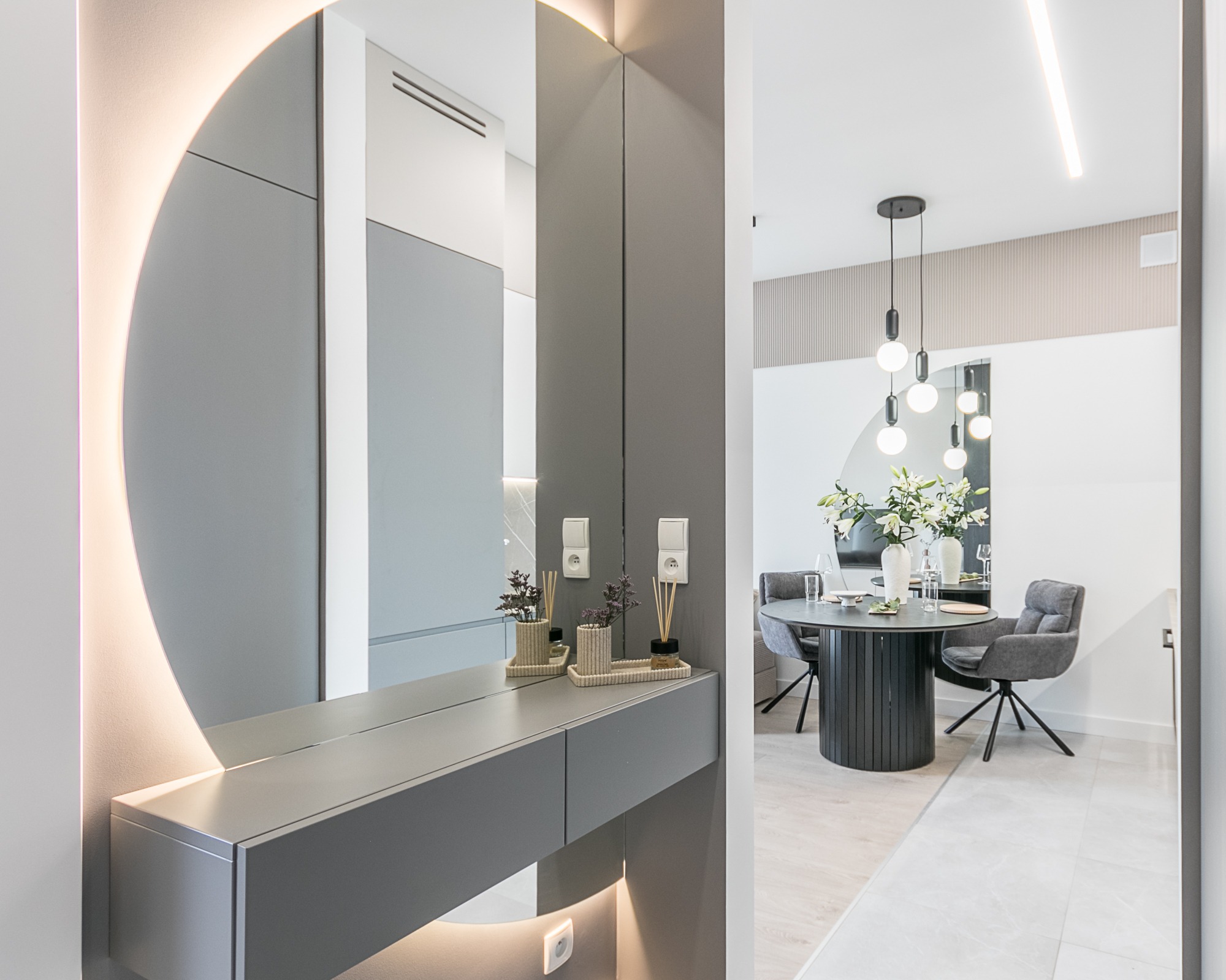 Nowoczesne wnętrze: szara konsola z okrągłym, podświetlanym lustrem i dekoracjami, odbicie stołu z kwiatami. Minimalistyczny design, subtelne oświetlenie.