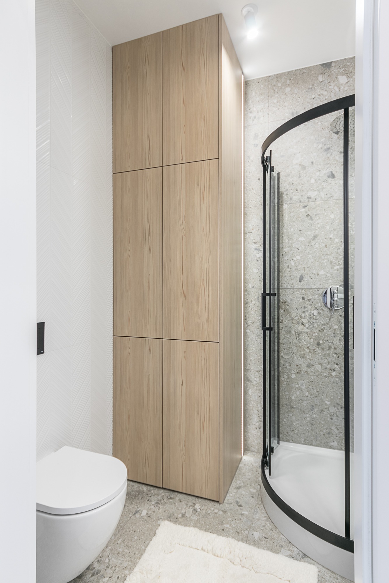 Nowoczesna łazienka z kabiną prysznicową z czarną ramą, drewnianą szafką, toaletą podwieszaną i mozaikową podłogą. Minimalistyczny design i jasna kolorystyka.