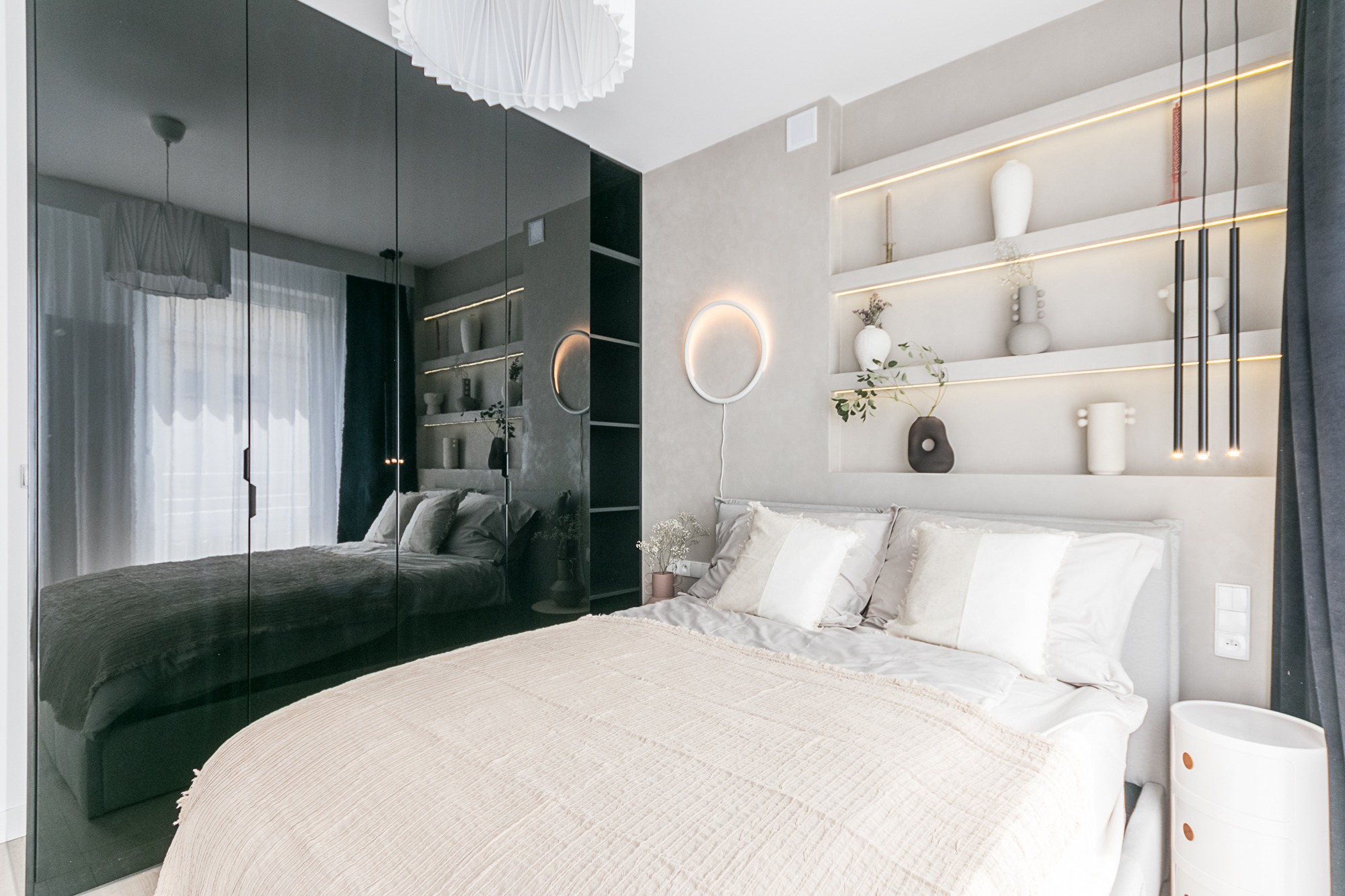 Nowoczesna sypialnia z lustrzaną szafą i podświetlanymi półkami. Jasne kolory, minimalistyczny design, okrągła lampa ścienna i beżowa narzuta na łóżku.