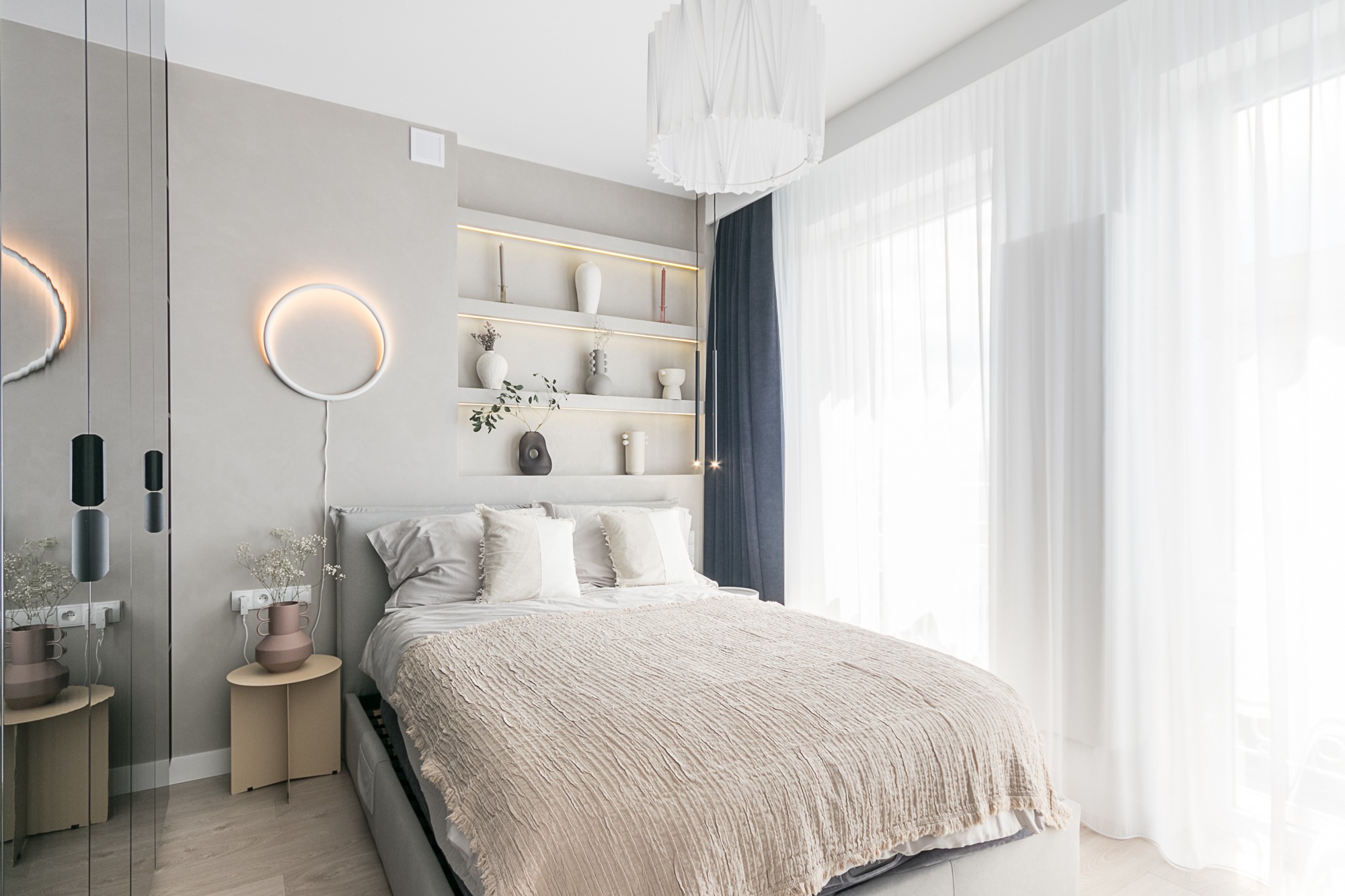 Jasna sypialnia z łóżkiem, okrągłą lampą na ścianie i półkami z dekoracjami. Minimalistyczny design z akcentami beżu i bieli. Duże okno z firanami.