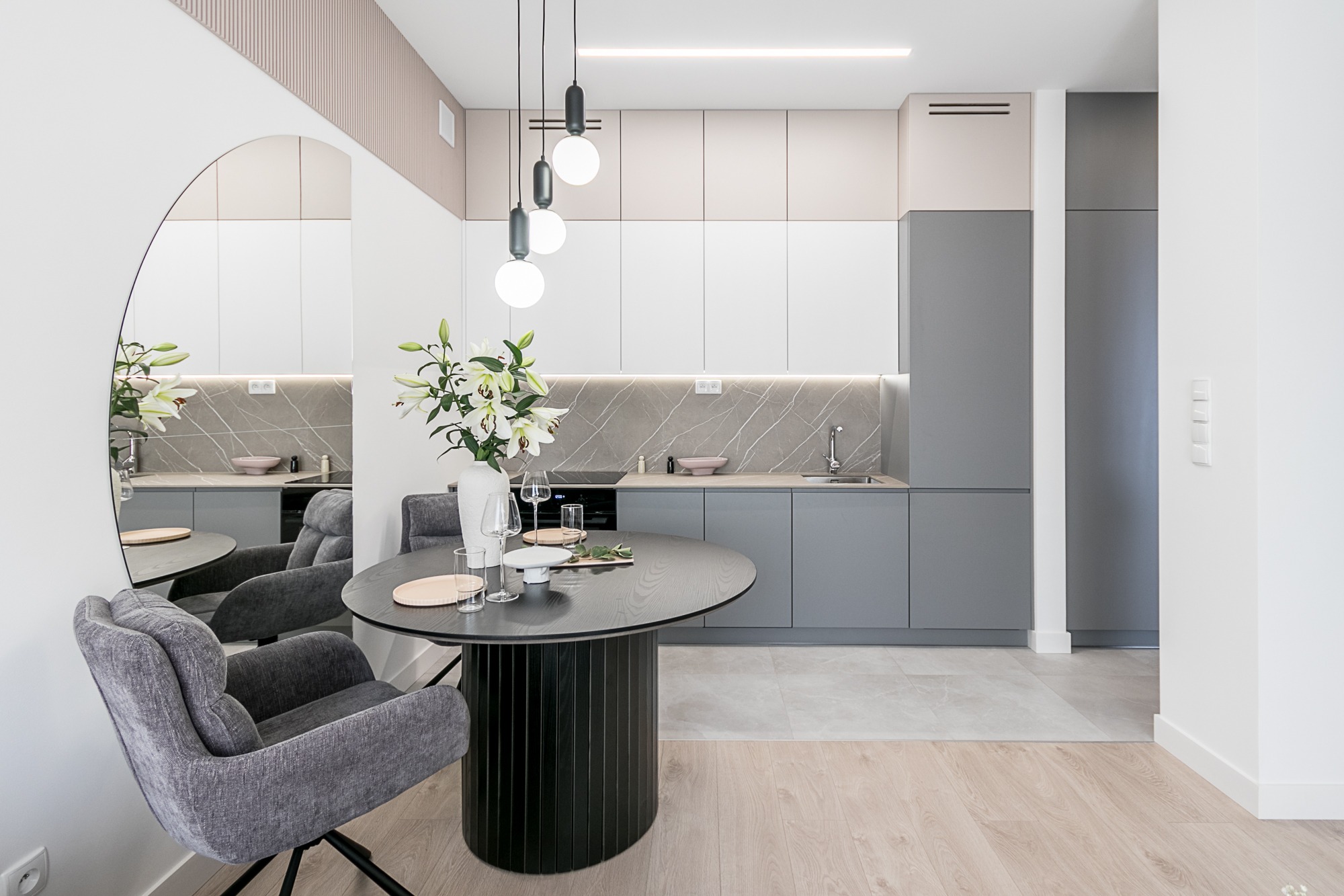 Stylowa aranżacja kuchni z okrągłym stołem i szarą zabudową. W tle marmurowy backsplash i nowoczesne oświetlenie. Całość w jasnych, stonowanych barwach.