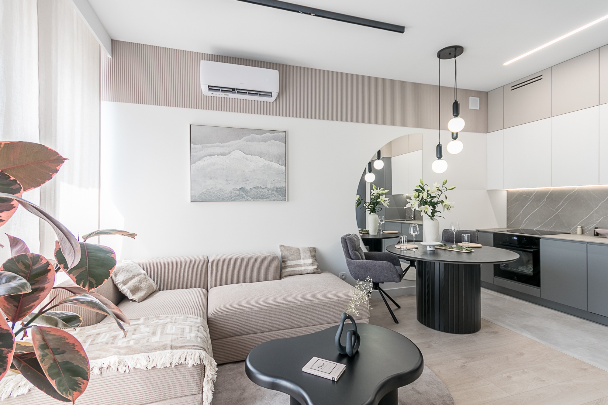 Jasne wnętrze z beżową sofą narożną, okrągłym stolikiem kawowym i jadalnią. Minimalistyczna kuchnia w odcieniach szarości i bieli. Całość dopełniają rośliny doniczkowe.
