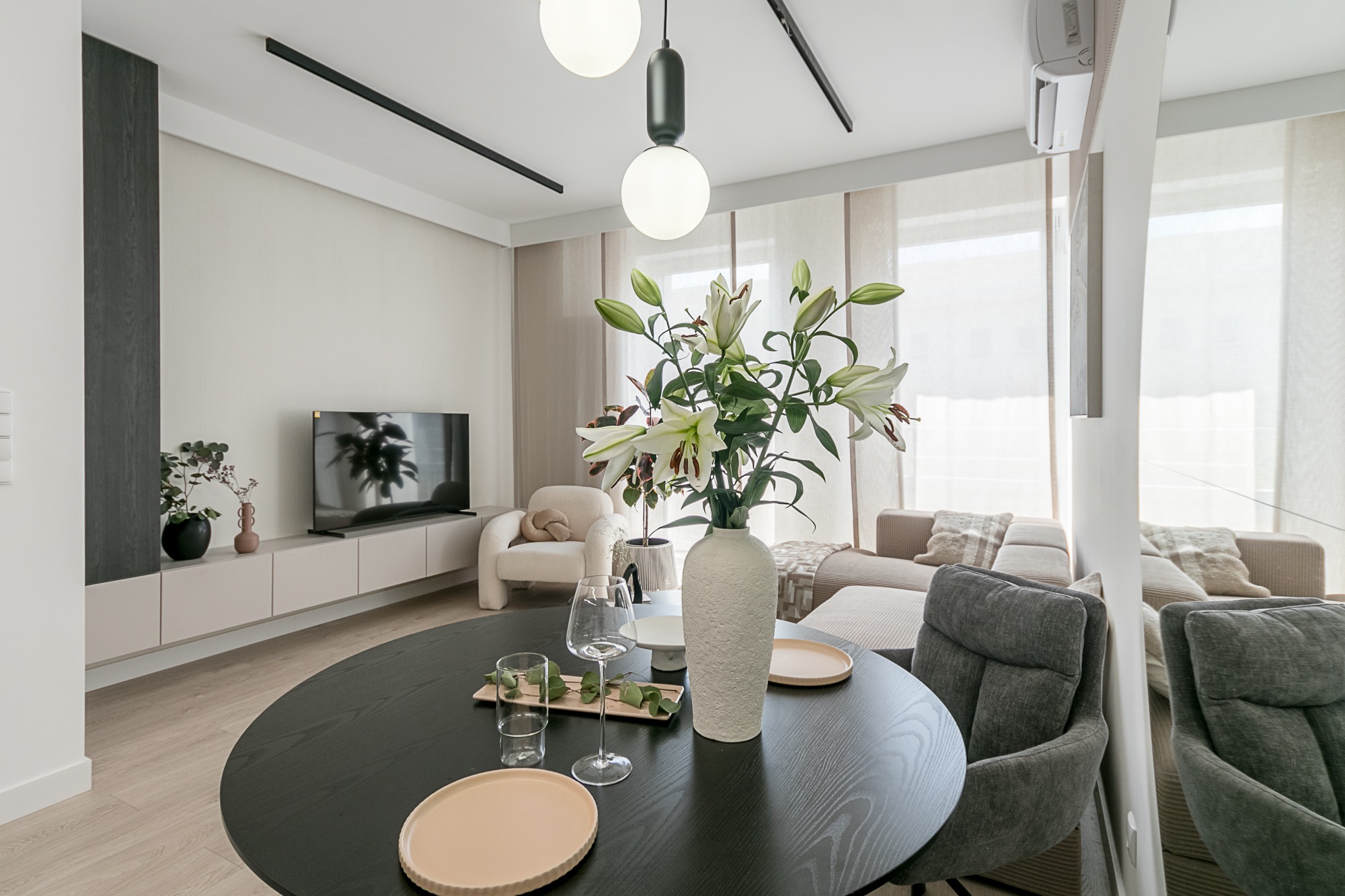 Stylowe wnętrze: okrągły, czarny stół z dekoracjami, wazon z liliami, szklanki, talerze. W tle jasne meble, telewizor i sofa. Minimalistyczny design z naturalnym światłem.