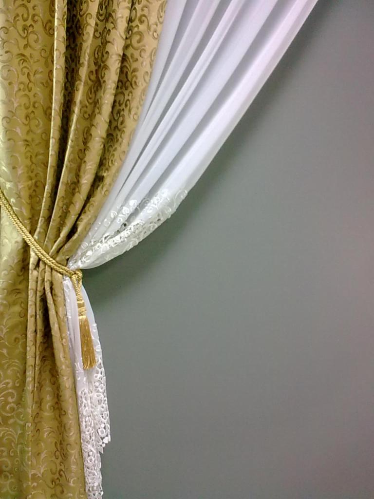 Upięta, złota zasłona dekoracyjna z motywem ornamentu, podpięta sznurem z frędzlami, z delikatną, białą firanką z koronkowym wykończeniem.