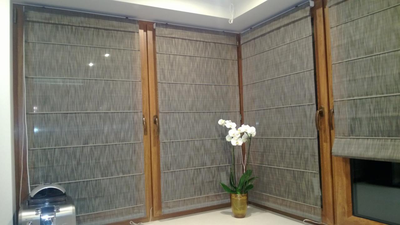 Zasłony rzymskie w odcieniach szarości zamontowane na drewnianych oknach, z białym storczykiem w doniczce w rogu pokoju.