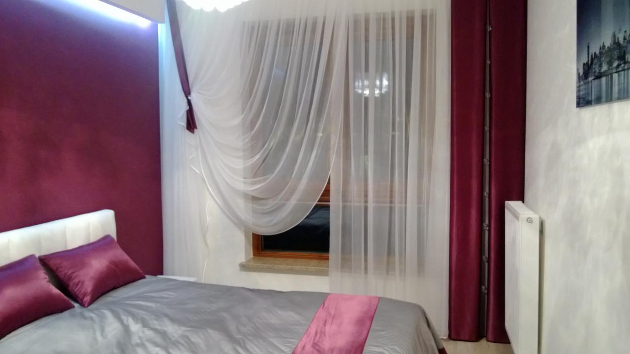 Sypialnia z zasłonami: białe, zwiewne firany i bordowe, ciężkie zasłony po bokach okna, szara pościel z bordowymi poduszkami i narzutą.