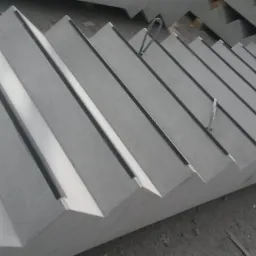 Stos prefabrykowanych, betonowych stopni schodowych z widocznymi elementami montażowymi, ułożonych jeden na drugim na szarym podłożu.