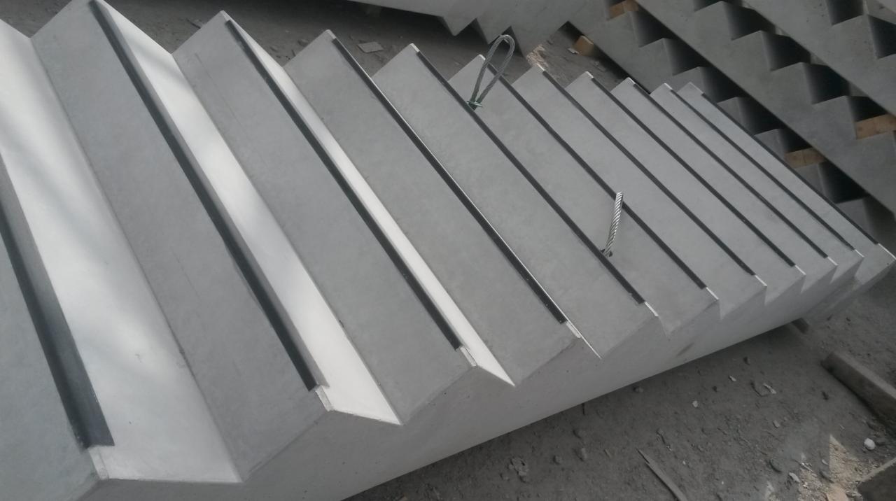 Stos prefabrykowanych, betonowych stopni schodowych z widocznymi elementami montażowymi, ułożonych jeden na drugim na szarym podłożu.
