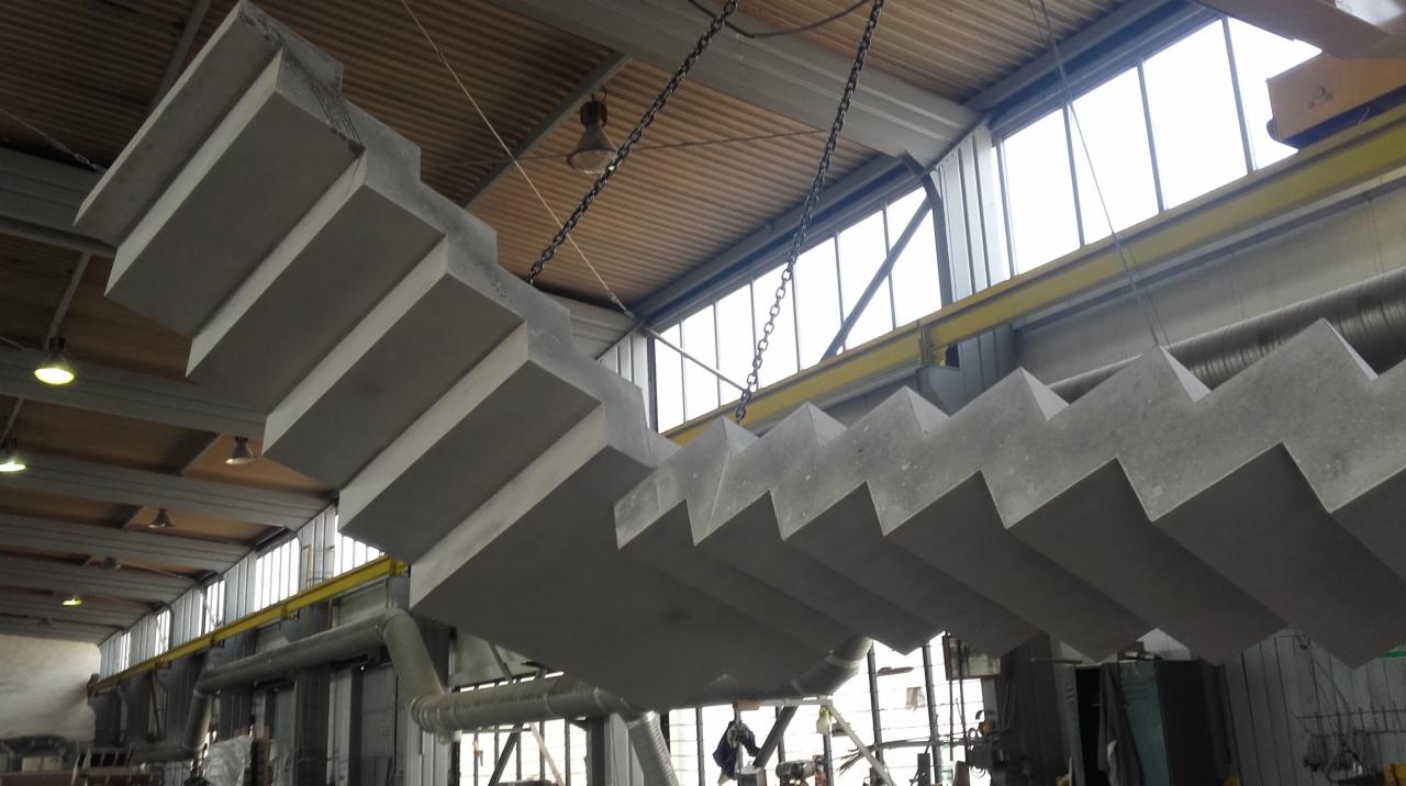 Prefabrykowane betonowe schody wiszące na łańcuchach w hali produkcyjnej, widoczne okna w tle.