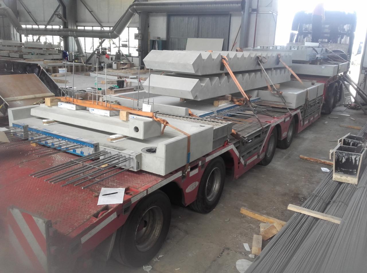 Ciężarówka z naczepą przewozi prefabrykowane elementy betonowe, zabezpieczone pasami transportowymi, widoczne wystające pręty zbrojeniowe i notatki na naczepie, w tle hala produkcyjna.