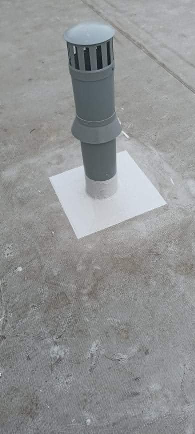 Szary kominek wentylacyjny na betonowym dachu, uszczelniony białą membraną hydroizolacyjną.