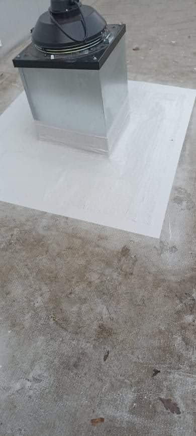 Metalowy komin wentylacyjny na płaskim dachu, uszczelniony białą, prawdopodobnie żywiczną, powłoką hydroizolacyjną na szarej powierzchni dachu.