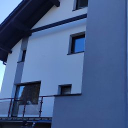 JORG JERZY PROKOPIUK - Pracownik na rusztowaniu maluje elewację nowoczesnego domu w odcieniach bieli i szarości, z balkonem ze szklaną balustradą, pod czystym niebem.