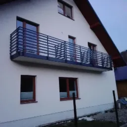 Nowoczesna, pozioma balustrada balkonowa z czarnych, metalowych profili na białym budynku z oknami w brązowych ramach, widok z dołu na tle zalesionego wzgórza.