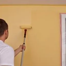 Mężczyzna maluje ścianę na żółto wałkiem, widoczny fragment okna i logo firmy na koszulce malarza.