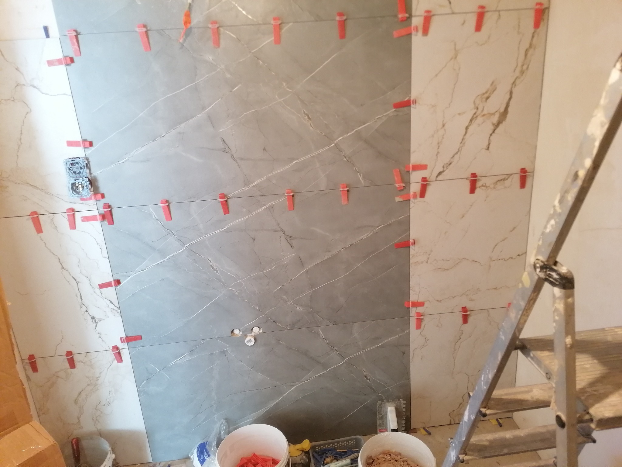 Układanie szarych i beżowych płytek imitujących marmur na ścianie, widoczne systemy poziomowania płytek, puszki elektryczne, drabina w tle.